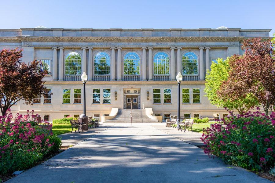 Photograph of the front of old Mesa County Courthouse in spring showing the wide staircase, four park benches, trees, flowering plants, shrubs, and grass area