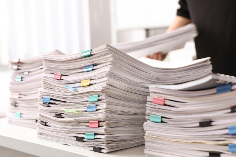 Photograph of three large piles of reports held together with binder clips on a desk with a man in the background with another report in his hands