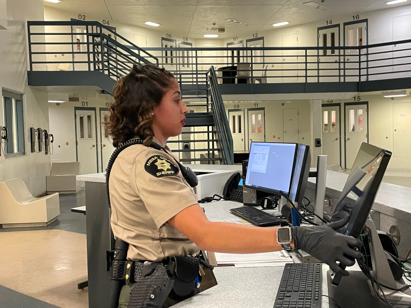 A female deputy stands at a counter touching a computer screen. Jail cell doors can be seen in the background.