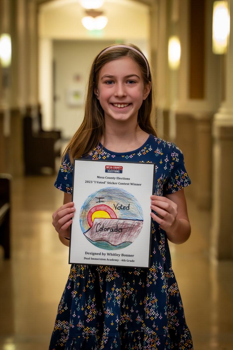 Elementary student holds up plaque with her winning "I Voted" Sticker Design. 