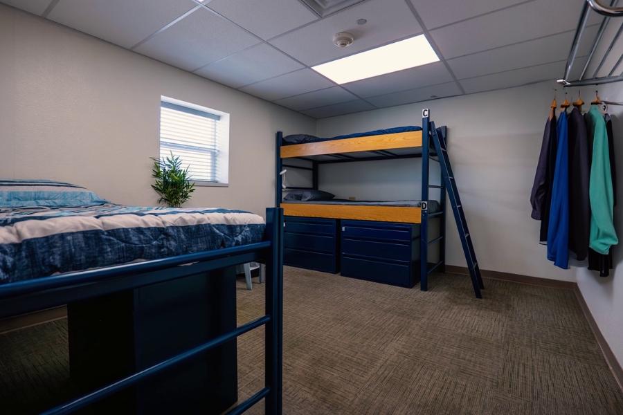 Dormitory with a window, a blue bunkbed, a blue single bed, and a nightstand with a plant on it.