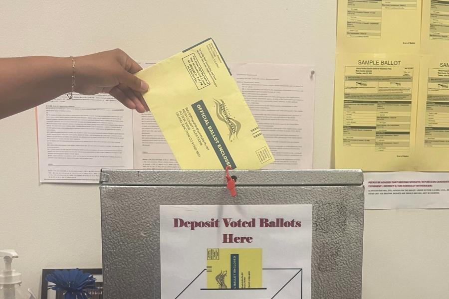 Hand putting yellow ballot into silver drop box. 